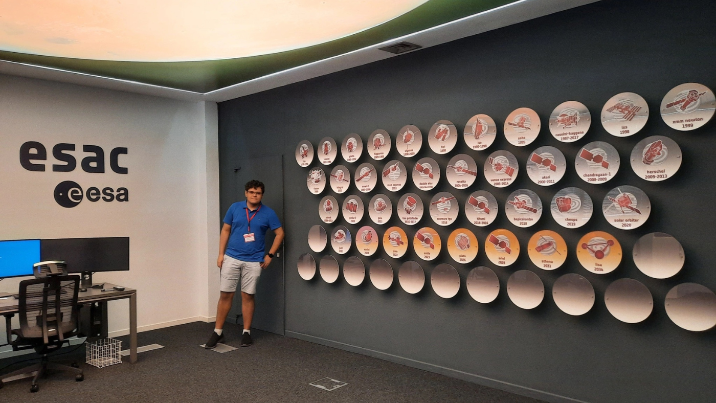 A person stands next to a wall display of circular plaques showcasing space missions at the European Space Astronomy Centre (ESAC). The ESAC and ESA logos are visible on the left above a desk with computer monitors