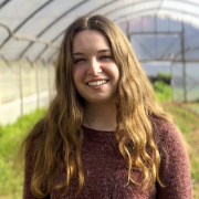 Smiling young woman with long hair in a maroon sweater stands in a greenhouse filled with plants.