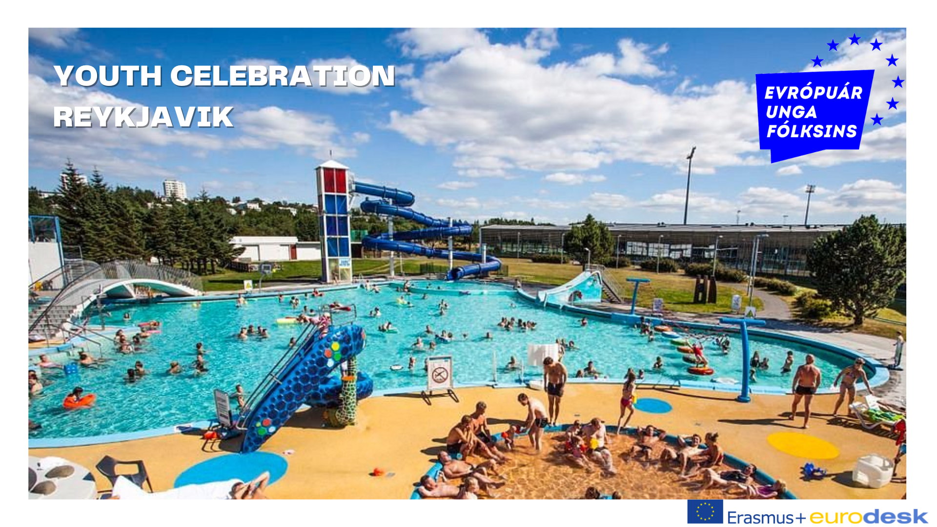 blue skies, white clouds and green trees in the back. Overview of a swimming pool on a sunny day. On the left side there is a bridge. On both sides of the pool there are three blue waterslides (one small near, two big further away). Many people enjoying the waters.