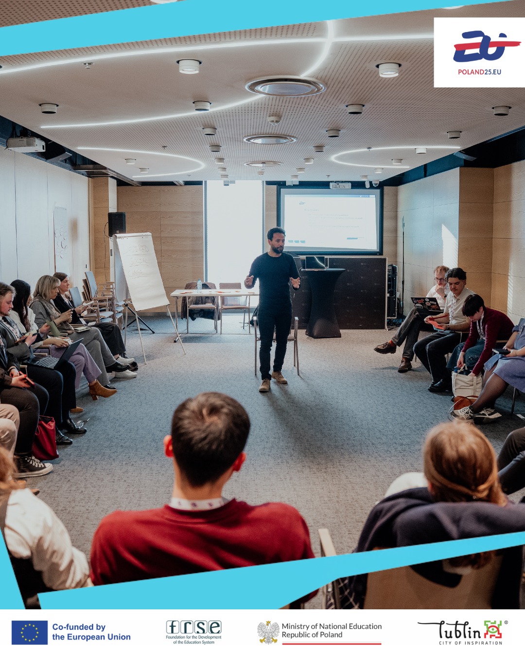 A man stands in the centre of a conference room, speaking to a group seated in a semi-circle. A presentation screen and flip chart are visible behind him. The room is well-lit with modern ceiling lights. Logos of Poland25.eu, the European Union, and other organisations are displayed.