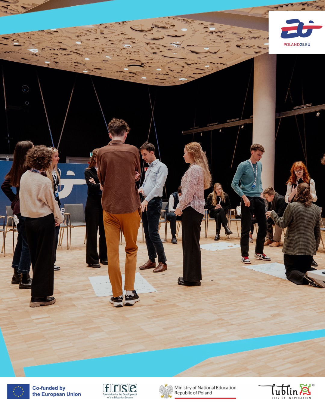 A group of people stands in a circle on a wooden floor, engaged in conversation. The room has a decorative ceiling and modern lighting. Logos for Poland25.eu, the European Union, and other organisations are displayed at the bottom.