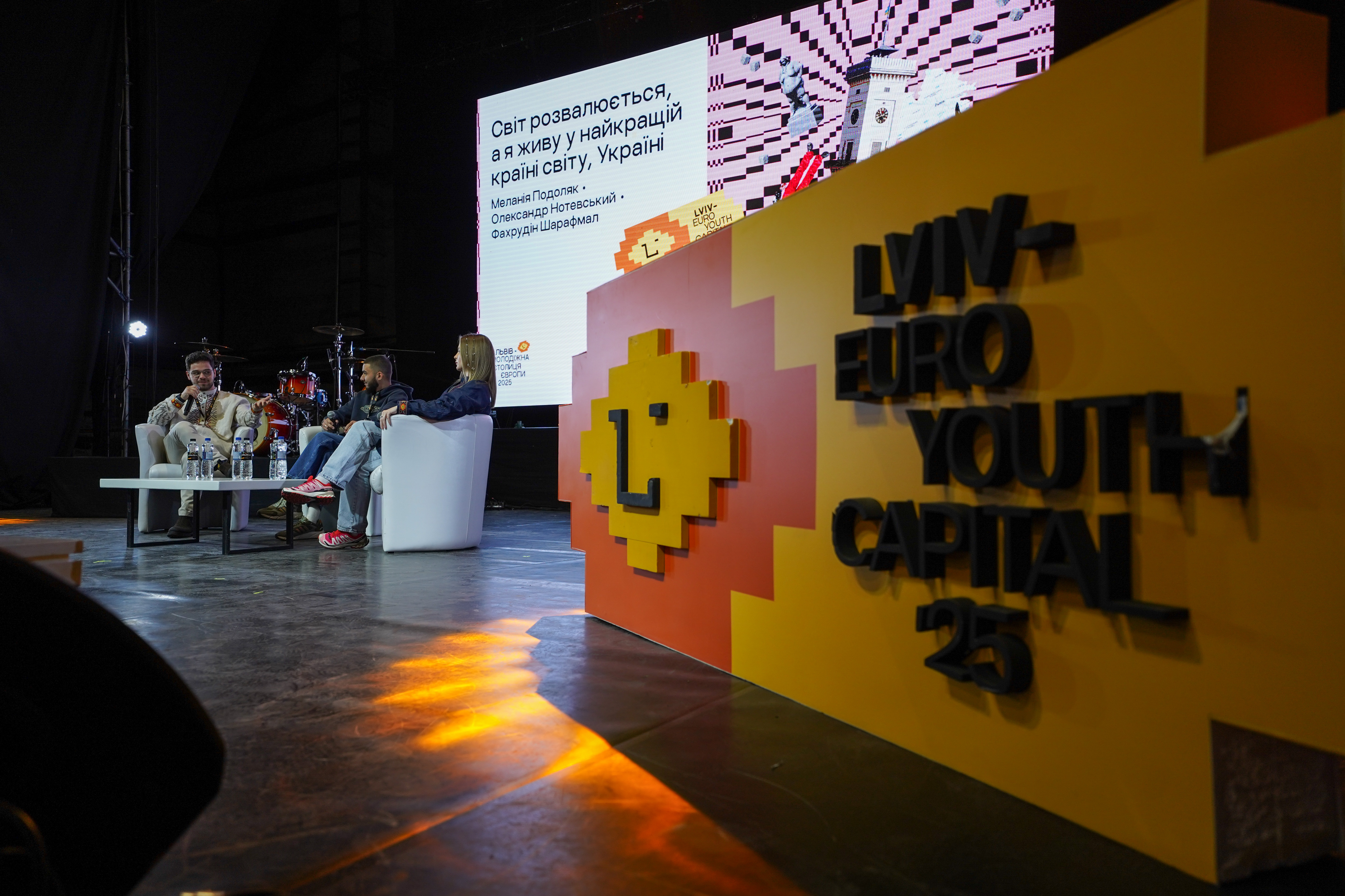 Three people on stage with a screen showing Ukrainian text. A yellow-red display reads “Lviv - Euro Youth Capital 25.” Drum set in the background, water bottles on table.