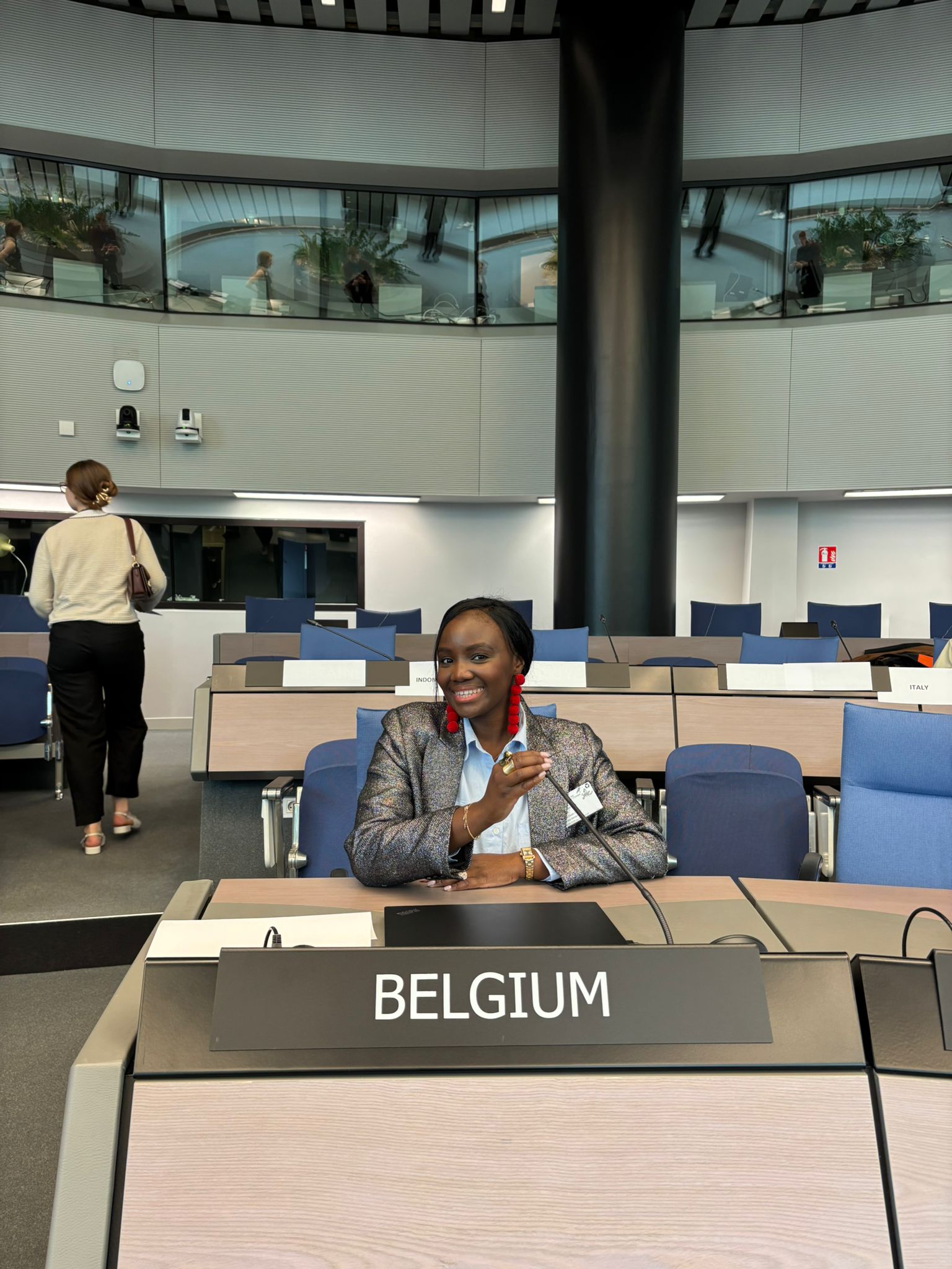 A person smiles, holding a microphone at a “Belgium” desk in a contemporary conference setting with empty chairs and upper level balconies.