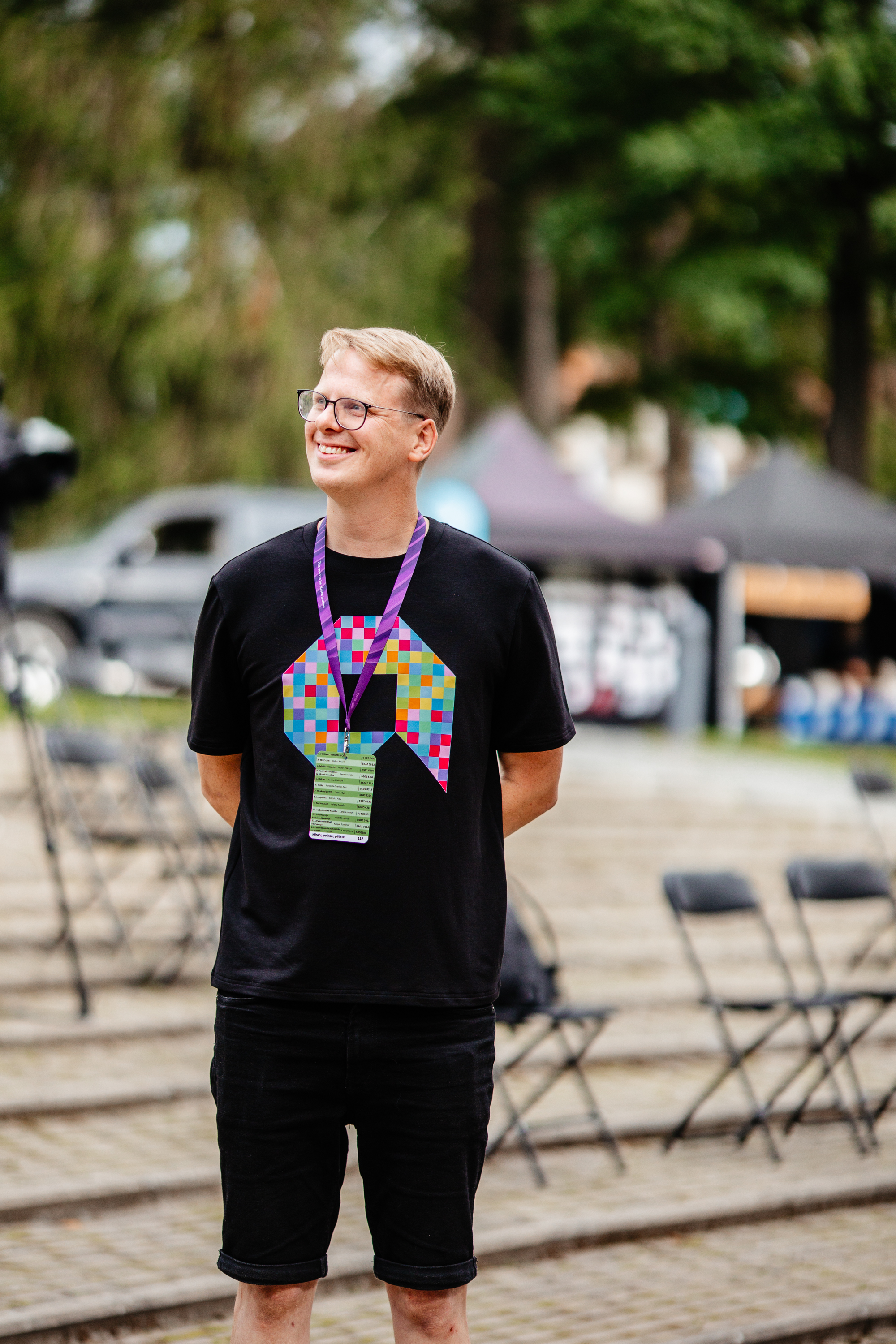 Smiling person in a black T-shirt with a colourful design, wearing a lanyard and badge, stands outdoors with chairs and tents in the background.