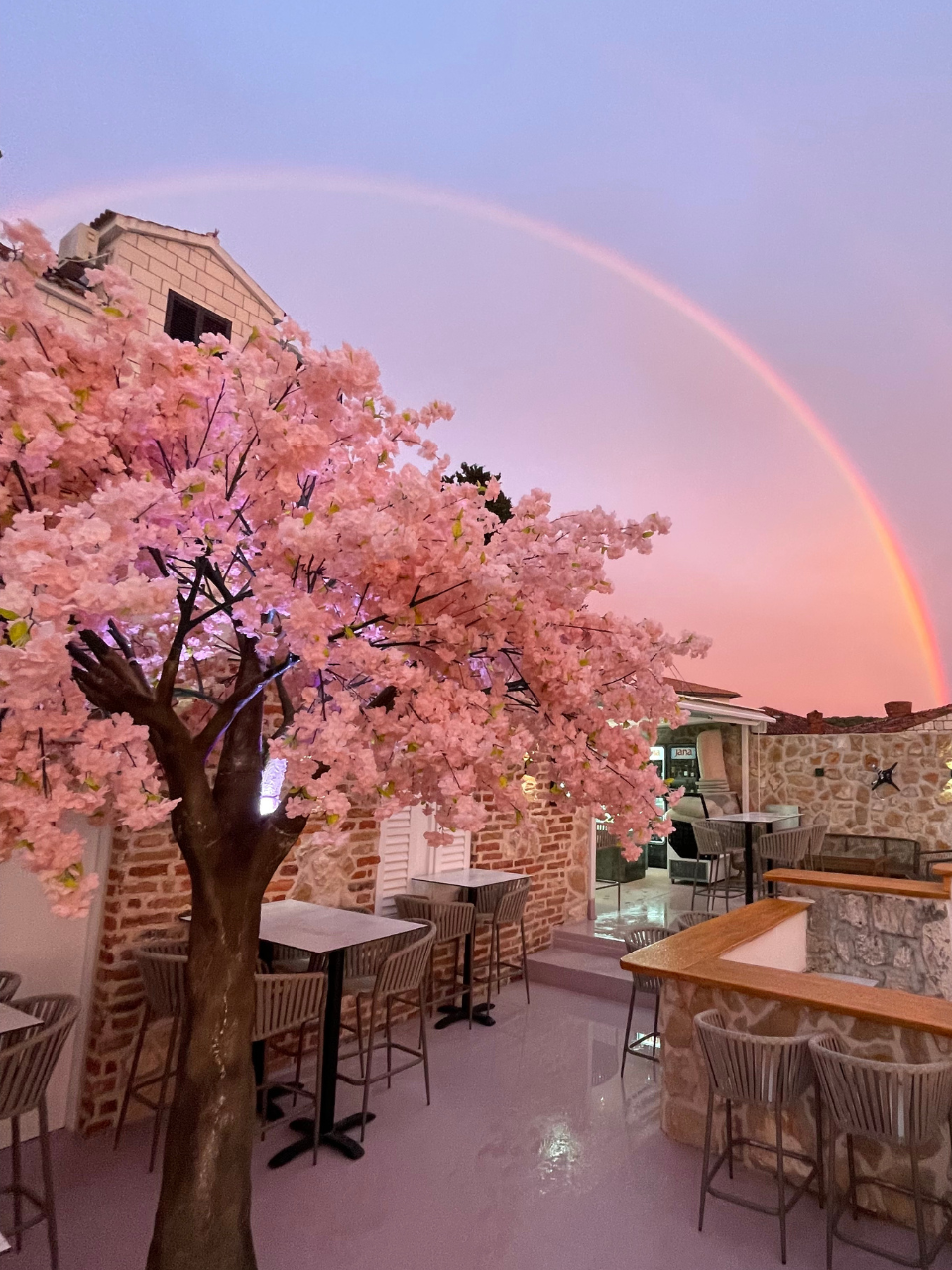 A charming outdoor café with a pink cherry blossom tree in full bloom. The setting sun casts a warm glow, highlighting a vibrant rainbow arching across the sky. Stone walls and tables with chairs create a cosy atmosphere. The scene is serene and picturesque.