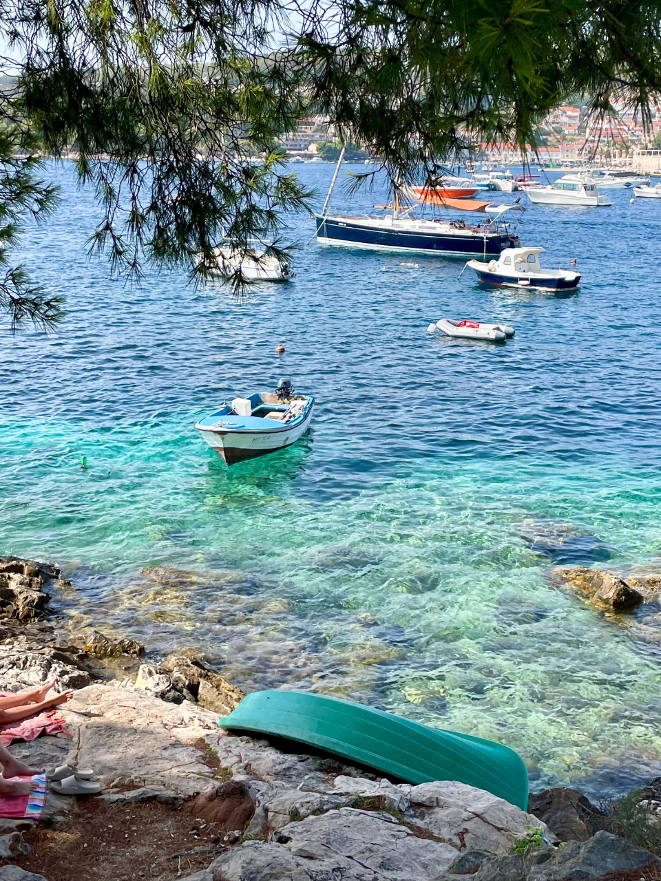 A serene coastal scene with clear turquoise water and several boats floating nearby. A small blue boat is closest to the rocky shore, where a green kayak rests. Pine branches frame the top of the image, and a person is sunbathing on a towel on the rocks. The background shows a distant town across the water.
