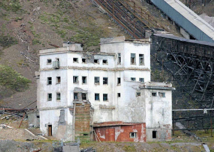 An old, abandoned building with peeling white paint and broken windows stands against a hillside. The structure is partially surrounded by rusted industrial equipment, adding to the sense of decay and neglect.