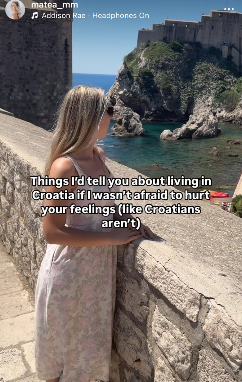 Woman in a dress stands by a stone wall, overlooking the sea and cliffs in Croatia, with an old fortress in the background
