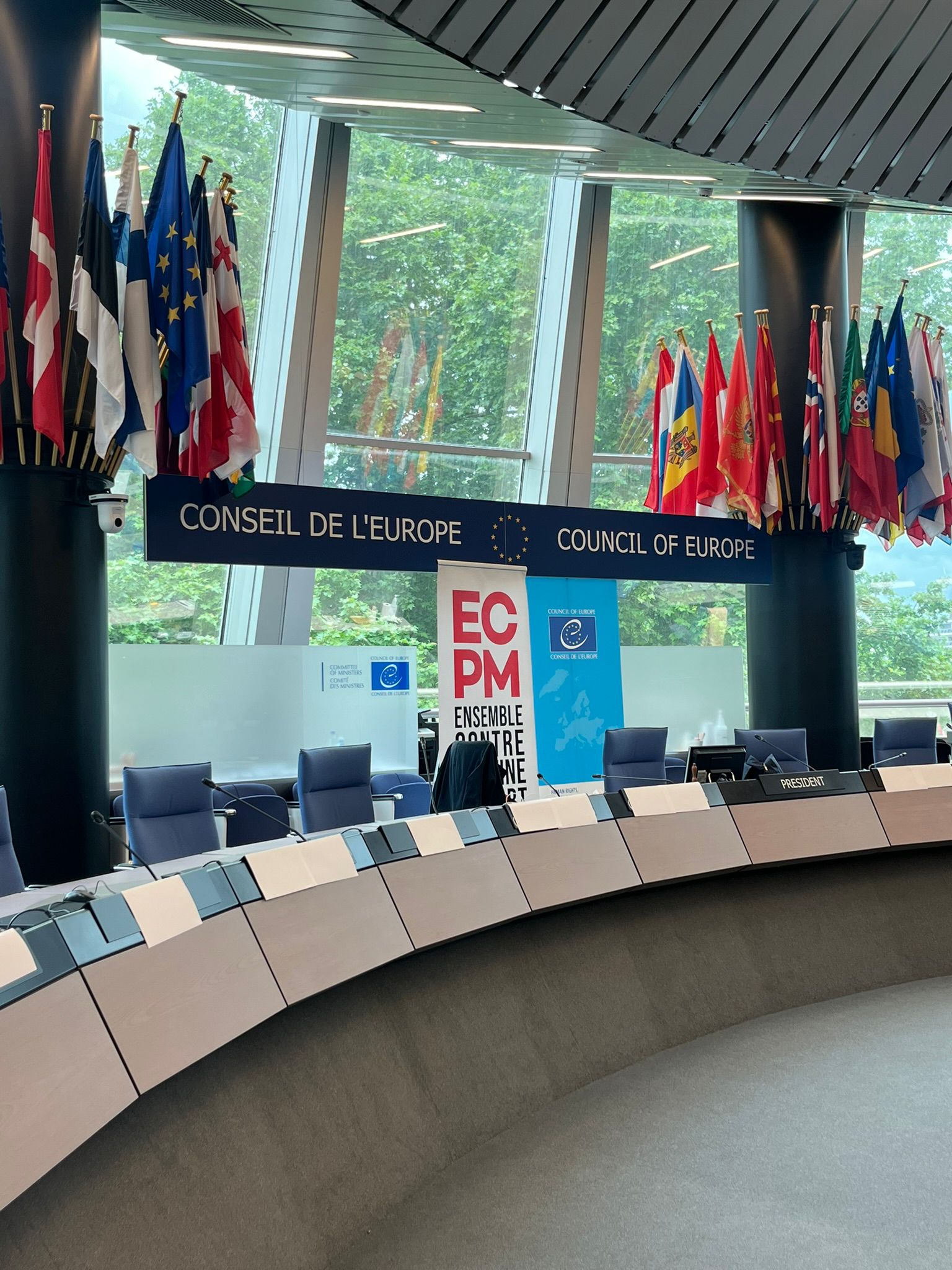 Conference room with a “President” desk, flags, and banners reading “ECPM” and “CONSEIL DE L’EUROPE”, featuring large windows and blue chairs.