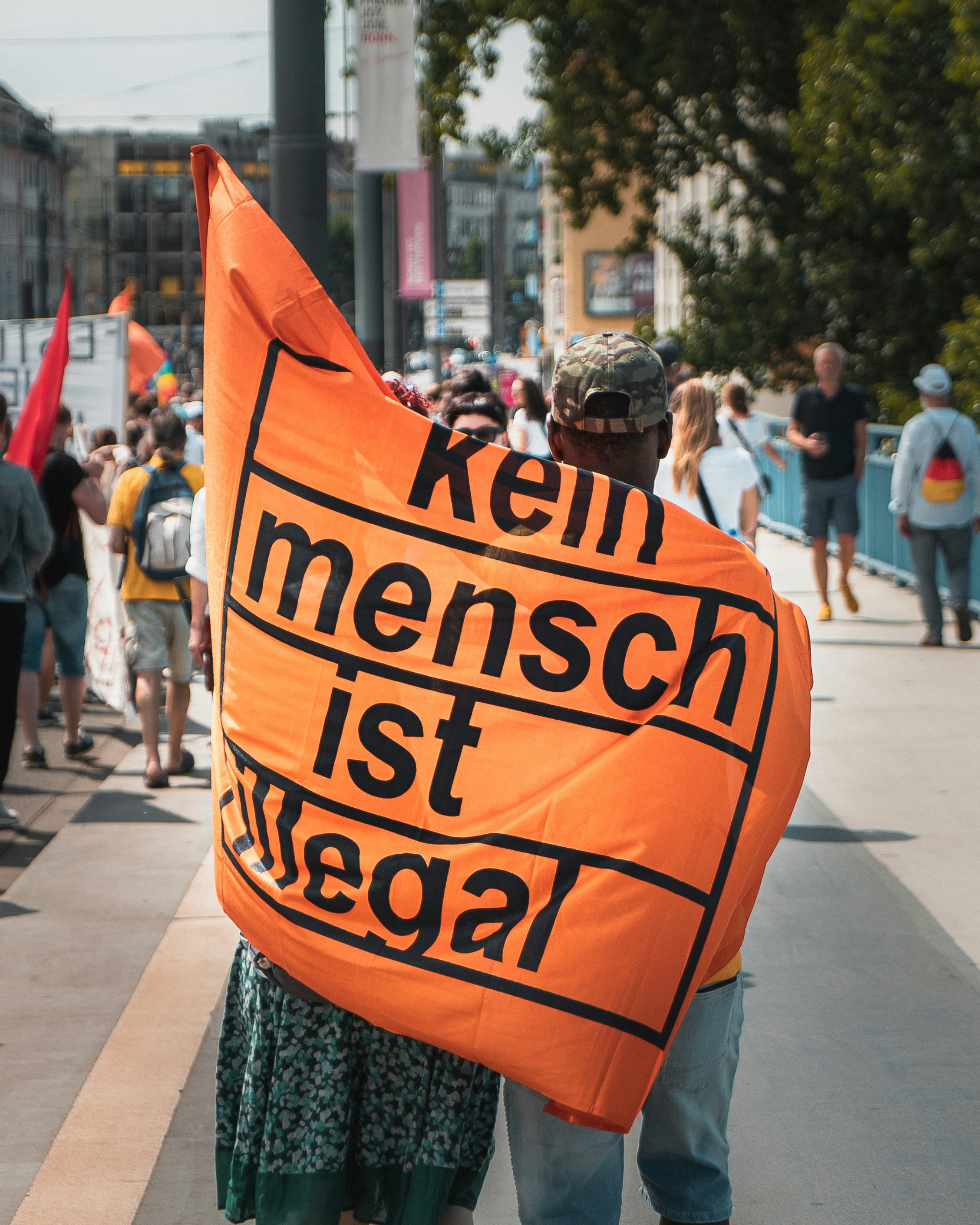 Two people at a protest hold an orange banner reading "Kein Mensch ist illegal," with a crowd and flags in the background.