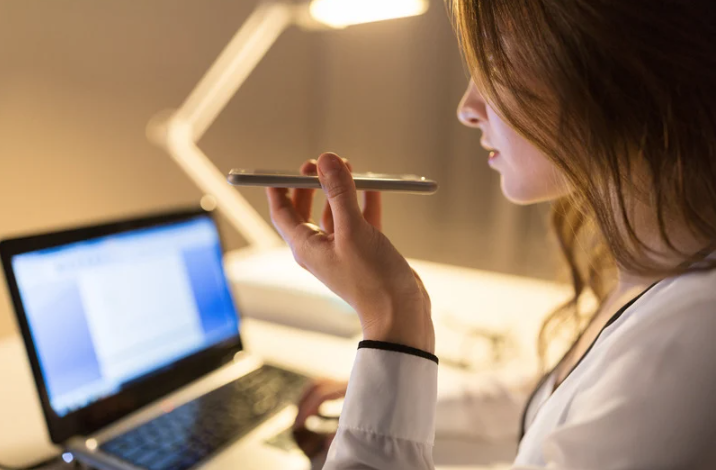 Close-up of a person using the voice recording function on her smartphone. In the background, you can see a laptop, a lamp all placed on top of a desk.