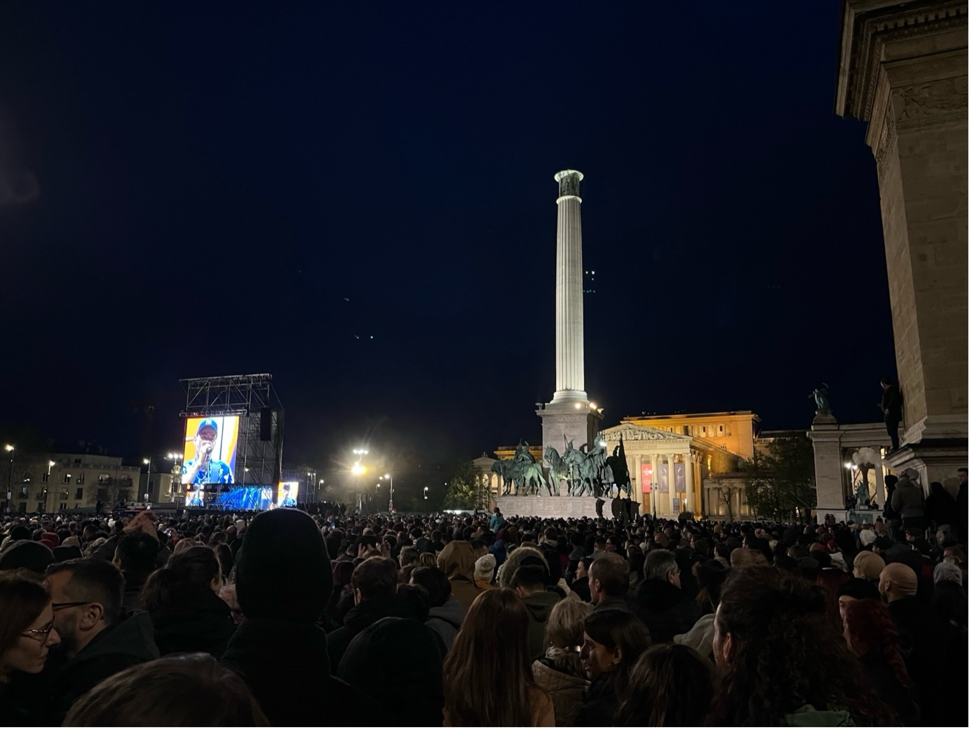 Nighttime crowd assembled in a square with a tall column and statues. A large screen shows a speaker, illuminated by lights. The scene is lively and bustling under a dark blue sky.