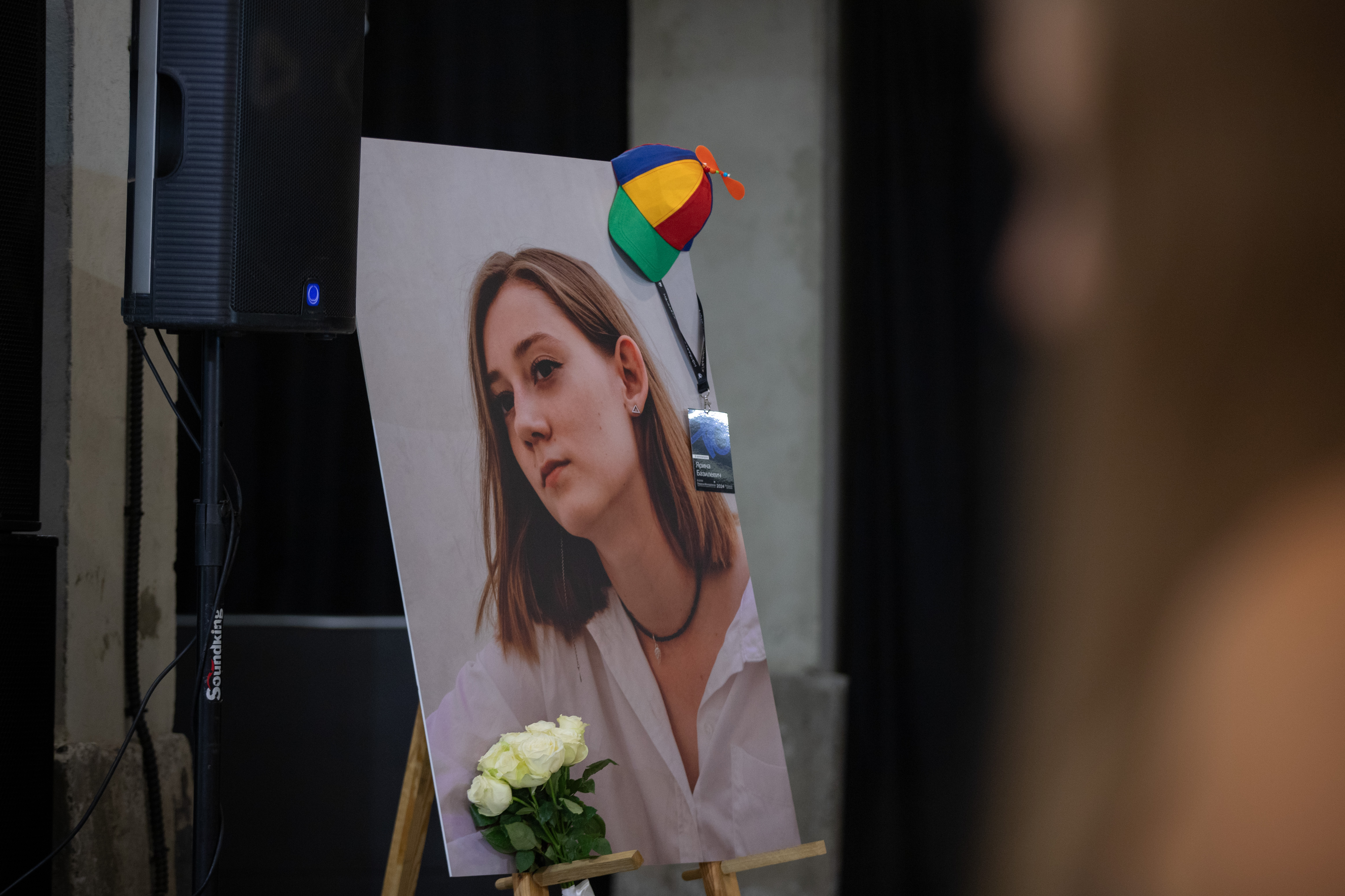 Young woman’s portrait on an easel with a propeller hat and name badge. White roses at the bottom. A speaker on the left and a blurred person in the foreground.