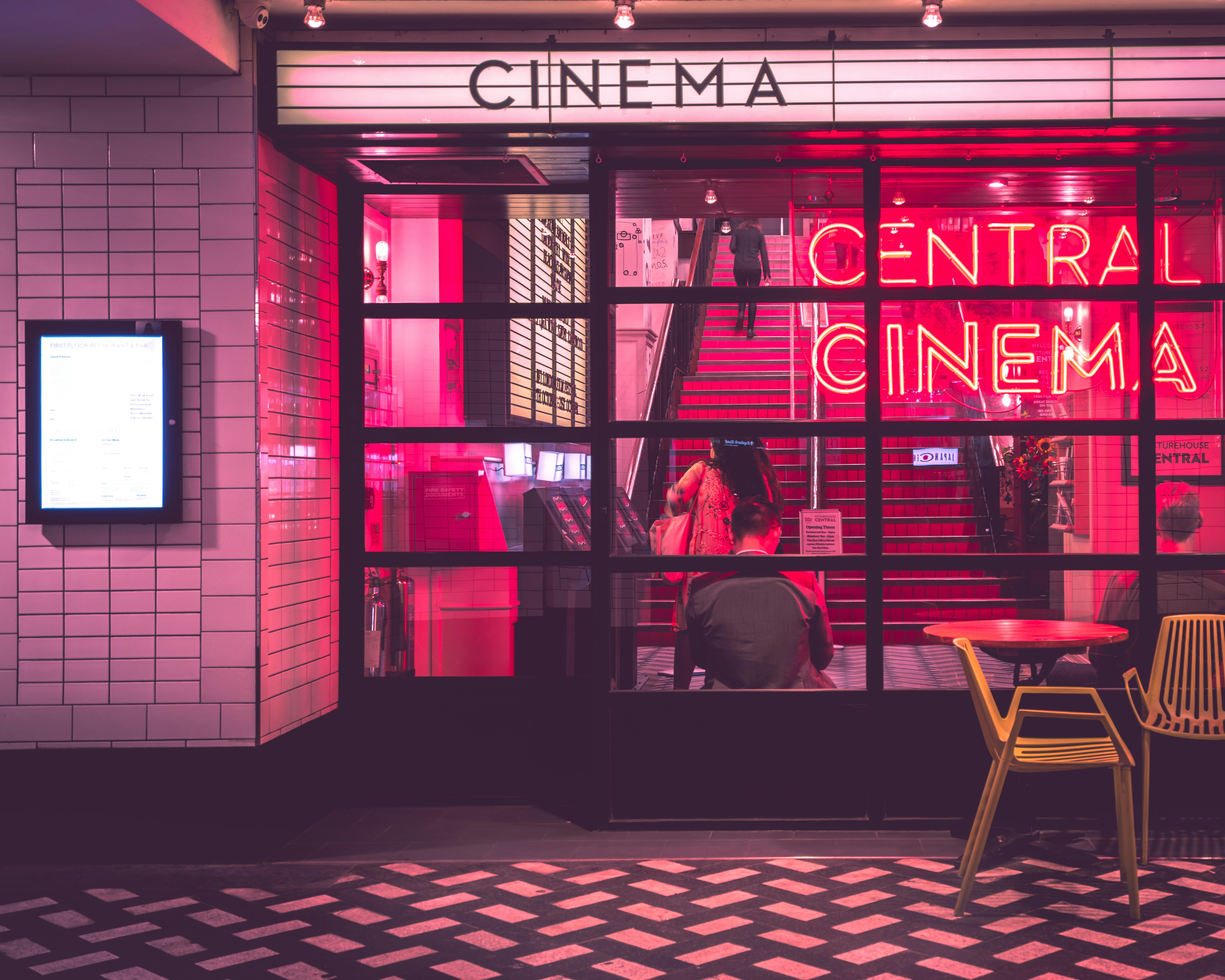 Entrance of a cinema with a neon “CENTRAL CINEMA” sign. There are people inside on a staircase bathed in red light. Outside, a man sits at a round table with a yellow chair, adding to the lively scene.