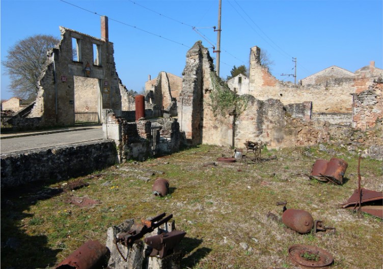 Ruins of a stone building with scattered metal remnants and overgrown grass under a clear sky.