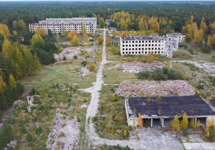 Aerial view of an abandoned area with dilapidated buildings surrounded by overgrown vegetation and trees. Rubble and debris are scattered across the site, indicating neglect and decay.