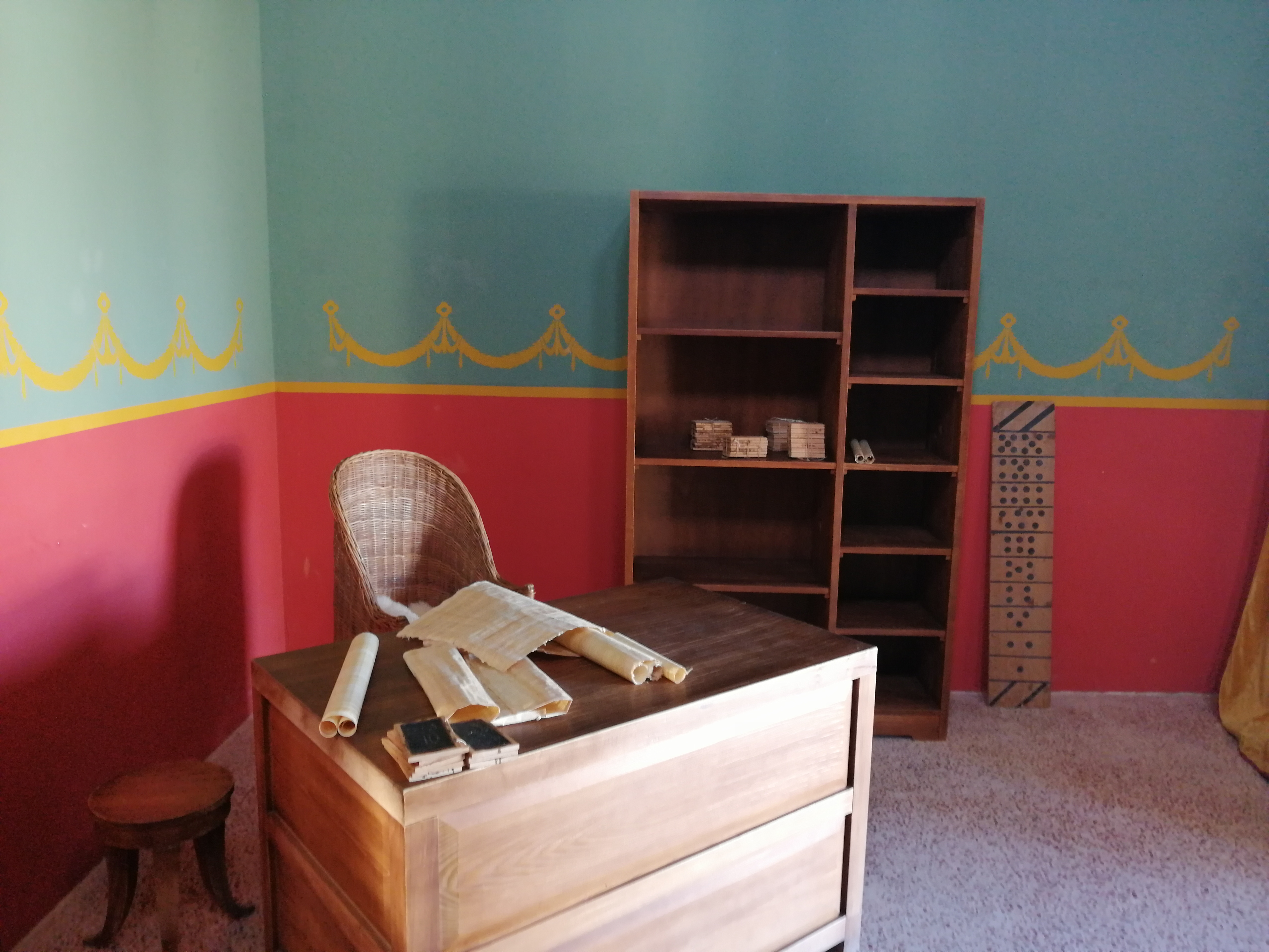A room with a wooden desk, scrolls, and a wicker chair. The teal and red walls have a yellow decorative pattern. A wooden shelf holds scrolls. A small stool is beside the desk.