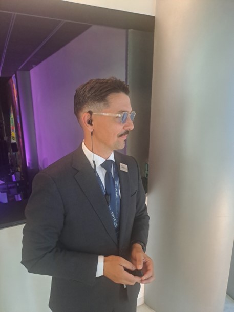 Man in a suit with glasses and earpiece in a modern, purple-lit space.