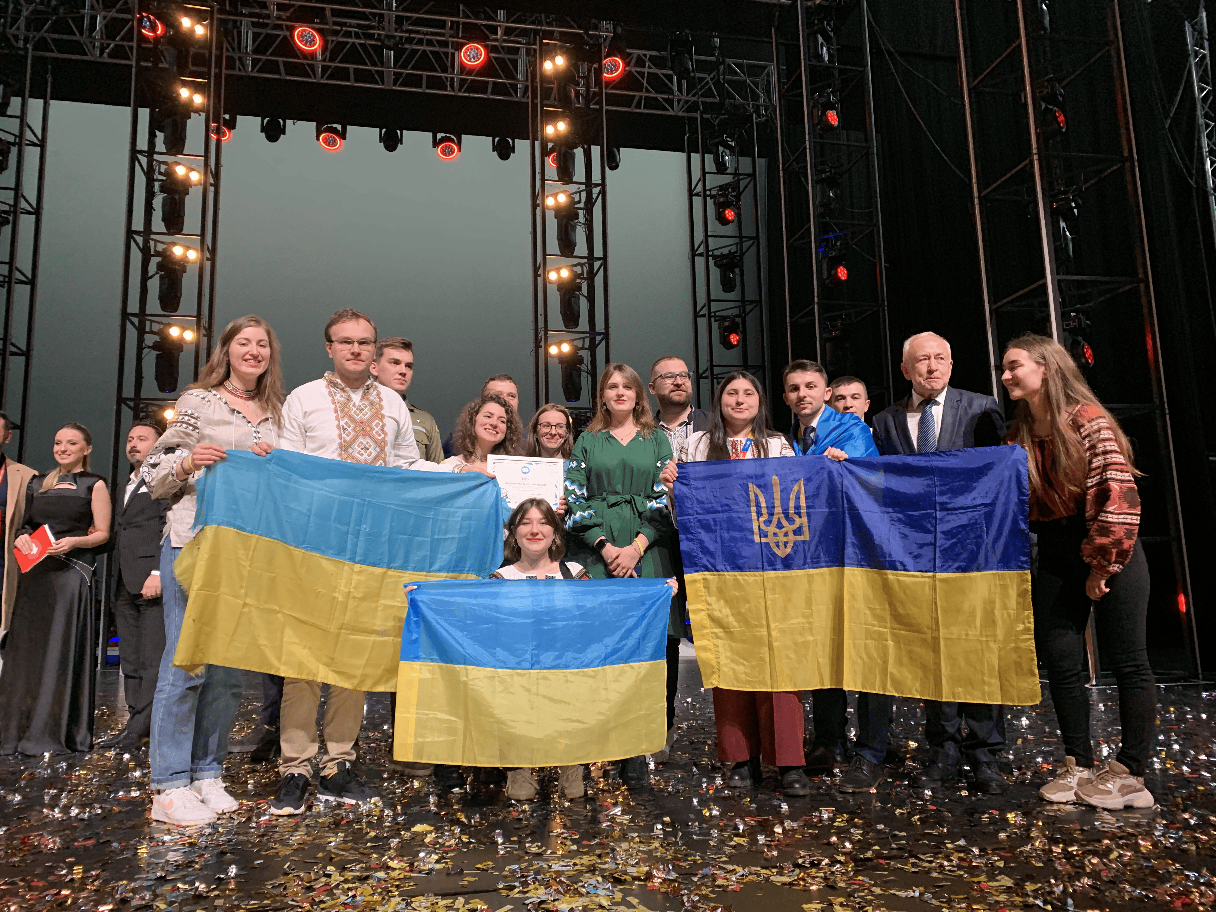 Group on stage with Ukrainian flags, including one with a trident emblem. Stage lights above and confetti on the floor. Some wear traditional embroidered outfits.