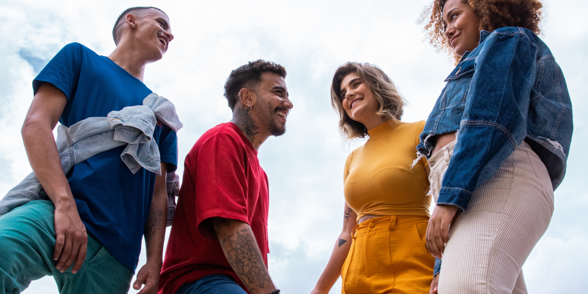 Four young adults stand outdoors, smiling and engaging with each other against a cloudy sky. They are dressed in casual clothing, including blue, red, yellow, and denim outfits.