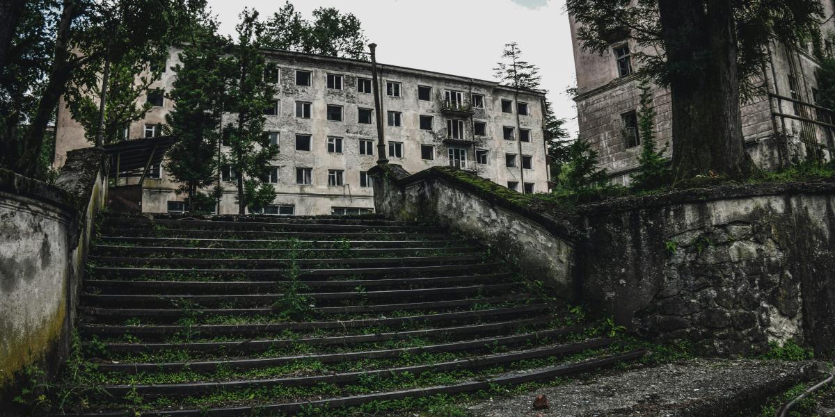 An abandoned stone staircase leads up to a dilapidated building with broken windows. The area is overgrown with greenery, and tall trees frame the scene, creating a sense of neglect and decay.