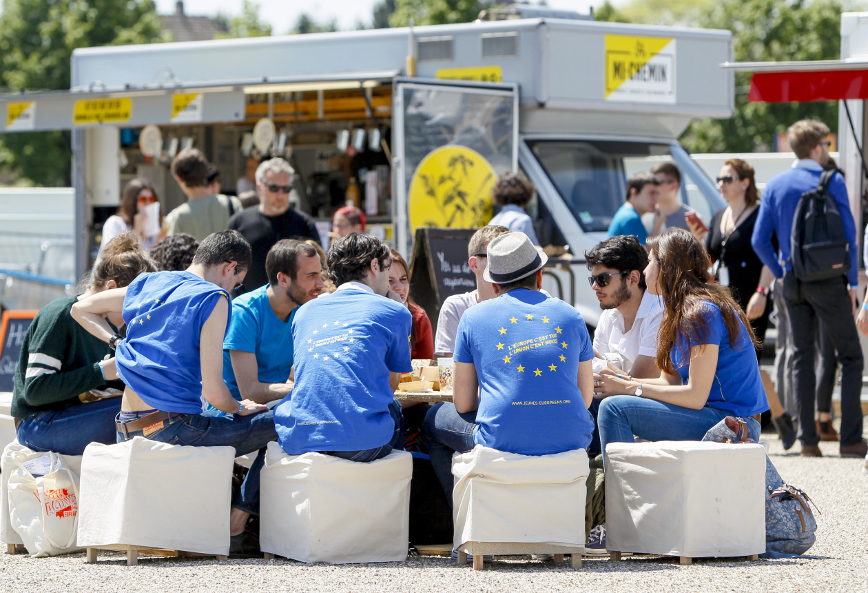 A group of young people sitting in a circle outside, eating and talking. They are wearing blue T-shirts with EU stars on their backs.