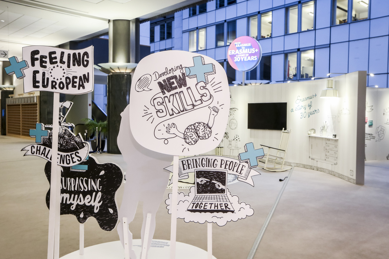 Exhibition space with white standees displaying motivational phrases and illustrations such as 'Developing New Skills' and 'Feeling European'. Background includes a large screen and a "Erasmus+ 30 Years" sign. Located in the European Parliament Brussels. 