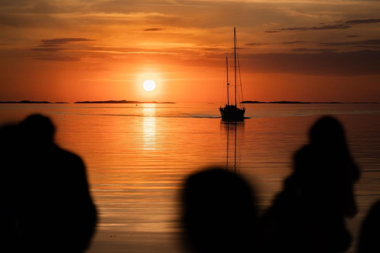 A sailboat on calm waters during a sunset, with an orange and yellow sky. Silhouettes of people are in the foreground.