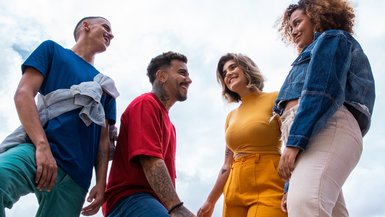 Four young adults stand outdoors, smiling and engaging with each other against a cloudy sky. They are dressed in casual clothing, including blue, red, yellow, and denim outfits.