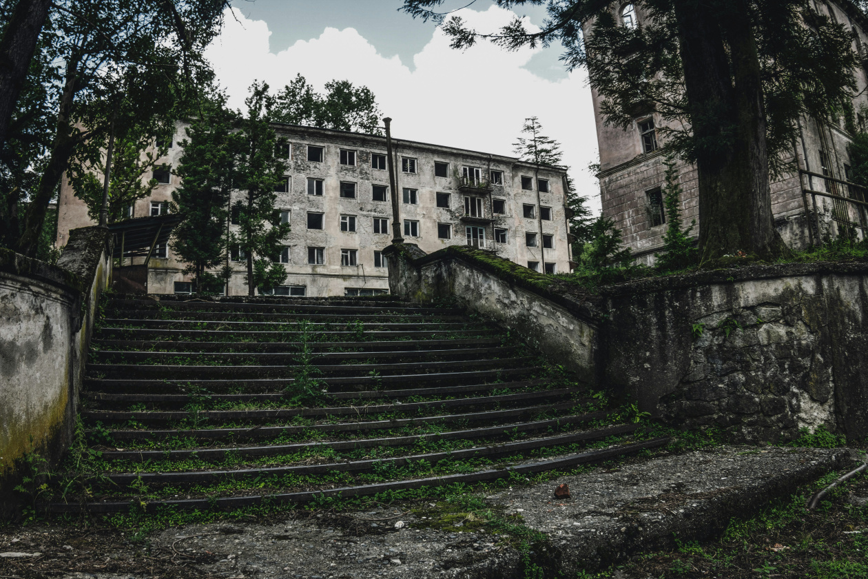 An abandoned stone staircase leads up to a dilapidated building with broken windows. The area is overgrown with greenery, and tall trees frame the scene, creating a sense of neglect and decay.