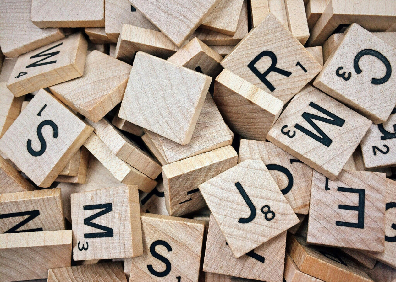 A pile of wooden Scrabble tiles with black letters and numbers on them, scattered randomly.
