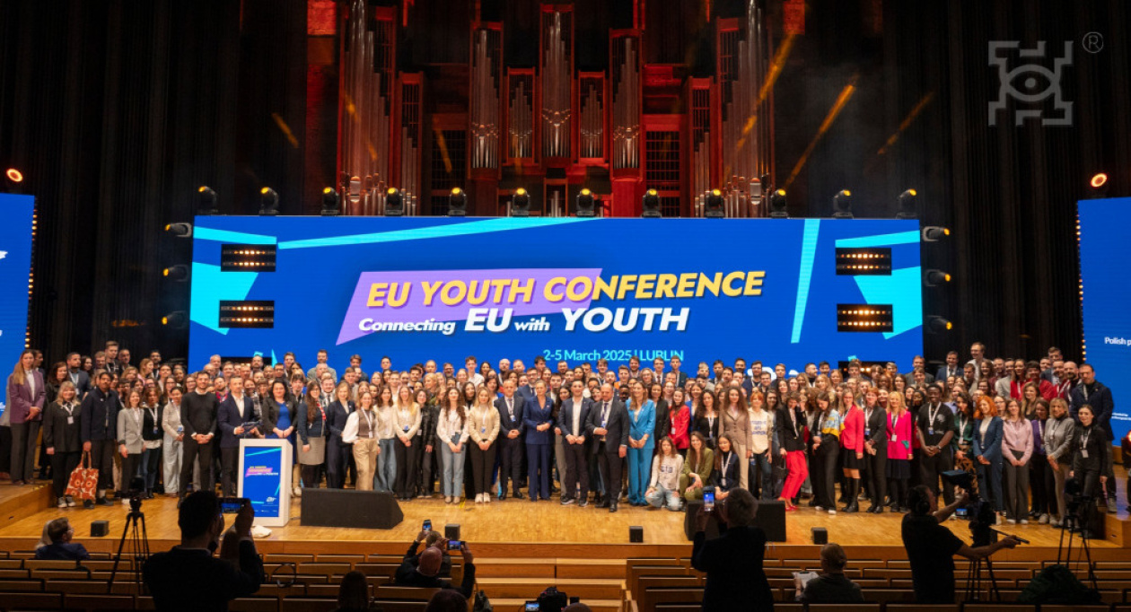 A large group of people stands on a stage in front of a big screen displaying "EU Youth Conference: Connecting EU with Youth, 2-5 March 2025, Lublin." The setting is a conference hall with an organ in the background. The attendees are diverse, dressed in formal and casual attire, and appear engaged.