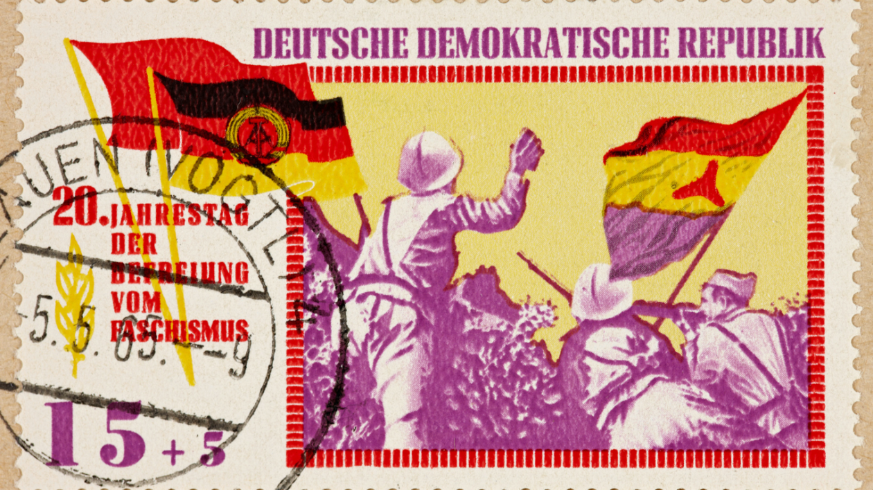 A postage stamp from the Deutsche Demokratische Republik features two soldiers raising flags. The background is yellow, and the soldiers are in purple. Text on the stamp commemorates the 20th anniversary of liberation from fascism. A postmark partially covers the image.