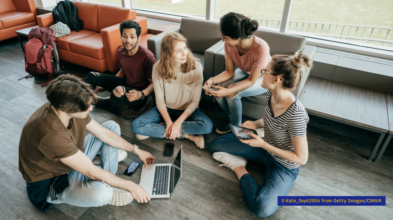 A group of young people, talking together and holding different technological devices, which include a tablet, a laptop and a smartphone.