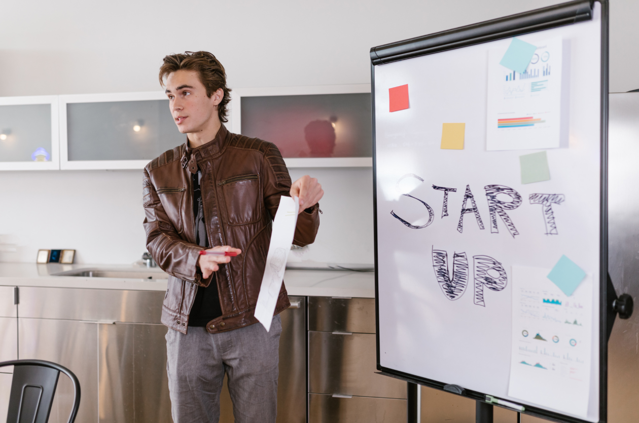 A man wearing a brown leather jacket is gesturing at a flip chart. The chart reads “START UP” with graphs and sticky notes attached. The scene appears to be in a modern office setting near a kitchenette.