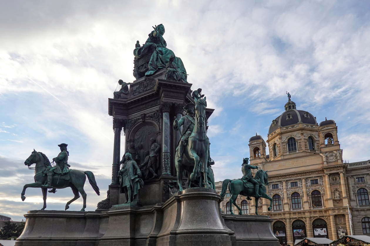 Monument with seated and equestrian statues in front of a domed neoclassical building under a cloudy sky.