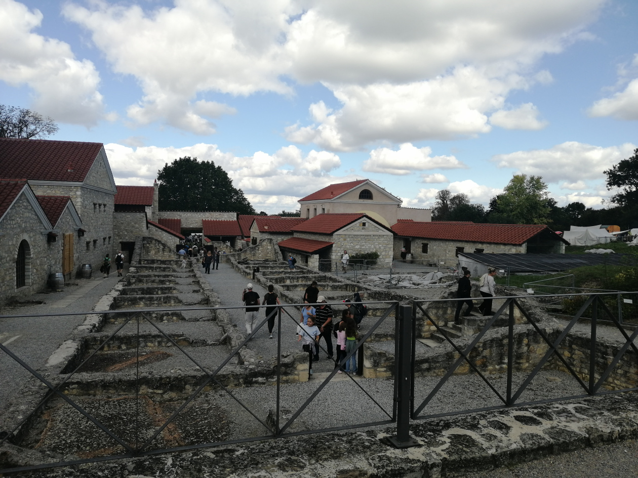 Visitors walk among ancient stone ruins and reconstructed buildings with red roofs at a historical site. Trees and a cloudy sky are in the background
