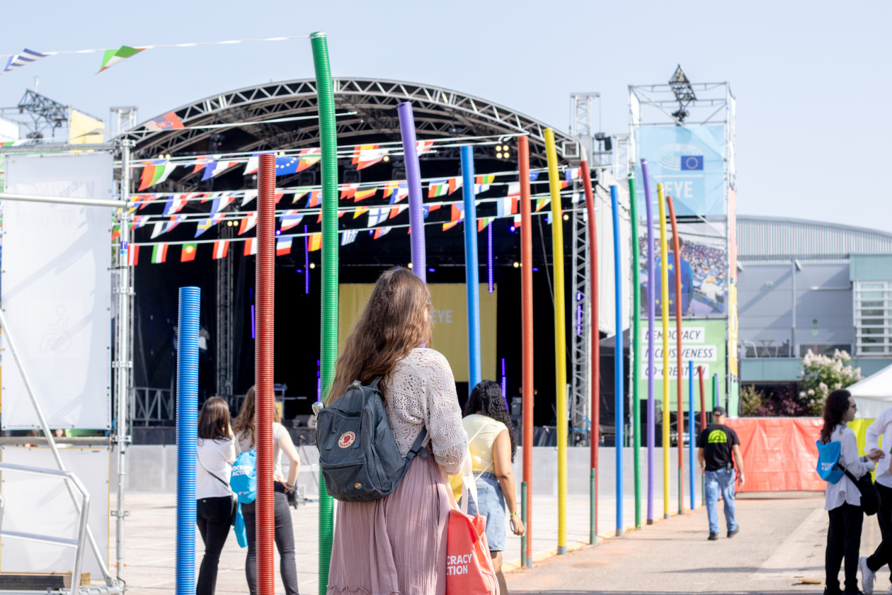 People walk towards a stage with flags and colourful poles, with “EYE” sign above.