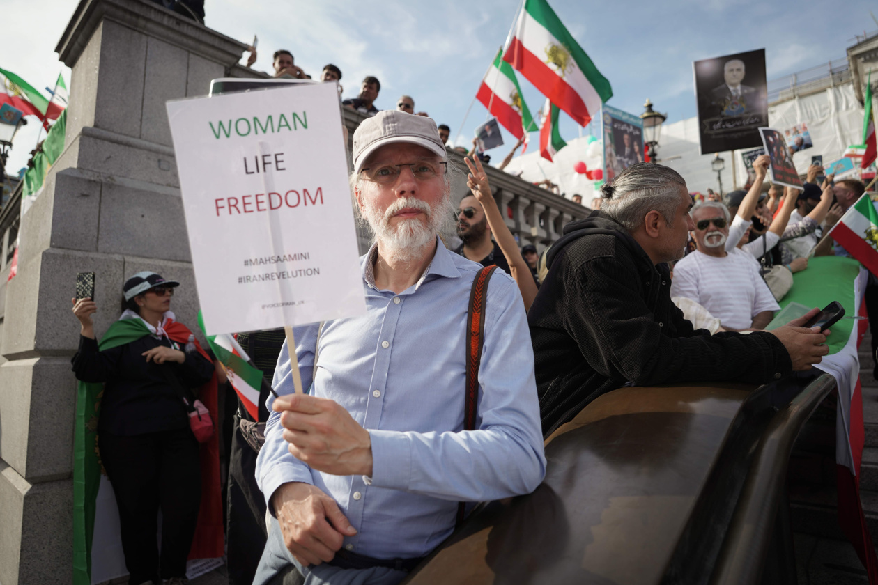 Man at protest holds “WOMAN LIFE FREEDOM” sign. Around him, people wave Iranian flags and display signs, creating a dynamic atmosphere.