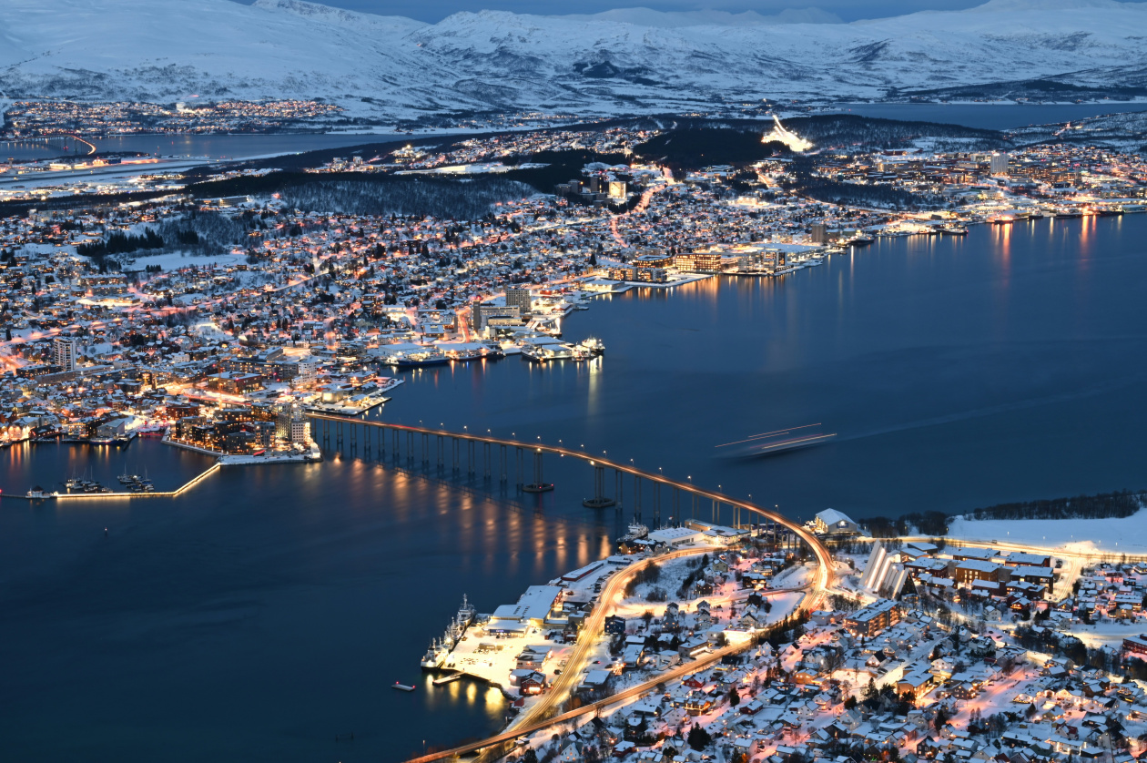 Snowy city by the sea at night, illuminated bridge over water, city lights reflecting, mountains in background.
