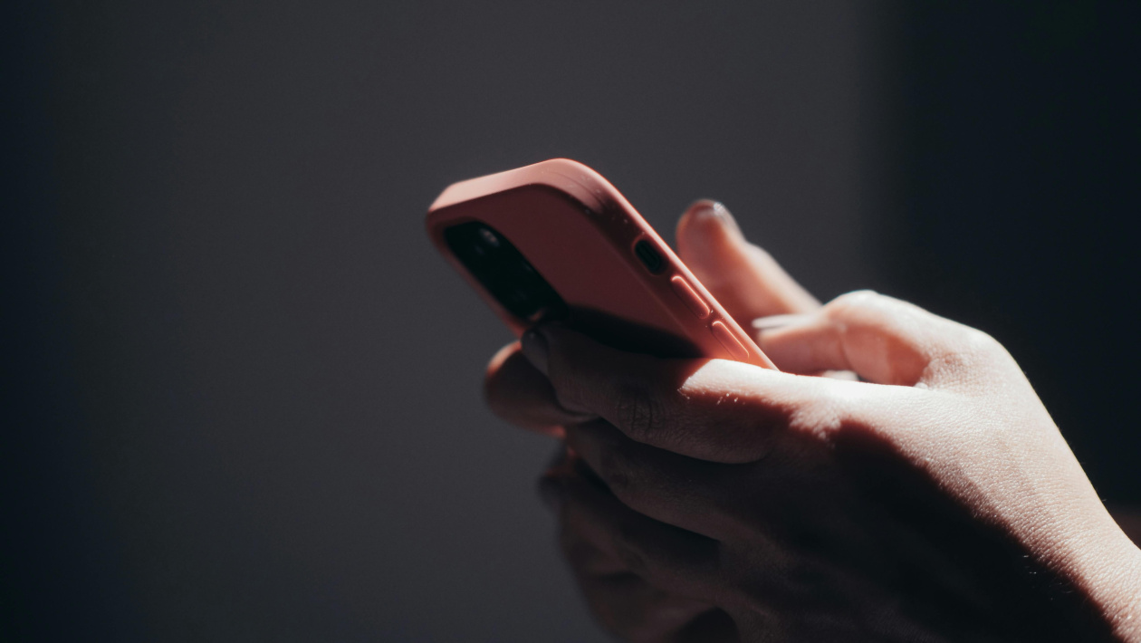 Hands holding a smartphone with a red case against a dark background, under focused lighting.