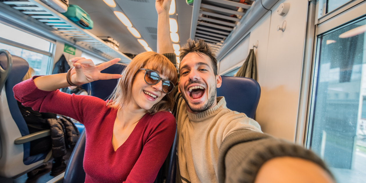 Couple taking a joyful selfie on a train, with the woman making a peace sign and both smiling broadly, surrounded by luggage and a scenic window view.