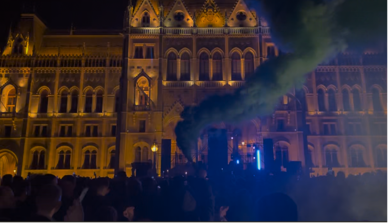 Nighttime crowd in front of a Gothic building with warm lights. Green smoke drifts through the scene, enhancing the atmosphere.