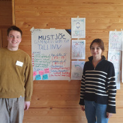 In the photo you can see boy and a girl standing in front of a wall. Boy is on the left and girl is on the right. On the wall inbetween them are some posters, where are written some ideas they got from the project writing camp. 