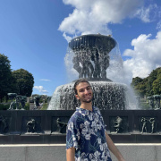 A smiling young man in a blue floral shirt stands in front of a large fountain with statues and cascading water. The sky is blue with scattered clouds, and trees are visible in the background.