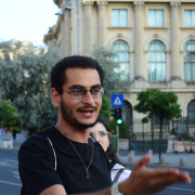 A young man with glasses and short curly hair speaks animatedly, gesturing with his hand. He wears a black t-shirt and a necklace. A historic building with arched windows and columns is in the background, along with trees and a pedestrian crossing sign.