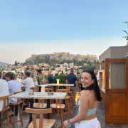 A woman in a light blue top and white skirt smiles with a shopping bag on a rooftop terrace. People are sitting at tables and the Acropolis is visible in the background under a clear sky.