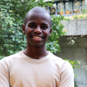 A person wearing a light-coloured sweater is smiling outdoors, with lush green trees and a building in the background.