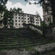 An abandoned stone staircase leads up to a dilapidated building with broken windows. The area is overgrown with greenery, and tall trees frame the scene, creating a sense of neglect and decay.