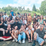 A group of young people are gathered outdoors, sitting and standing on a wooden platform and stone path. They are smiling and making heart shapes with their hands. The background features greenery and a building with a red roof. The atmosphere is cheerful and relaxed.  Alt-Text: A group of young people smiling and making heart shapes with their hands, sitting and standing outdoors on a wooden platform and stone path, with greenery and a building in the background.